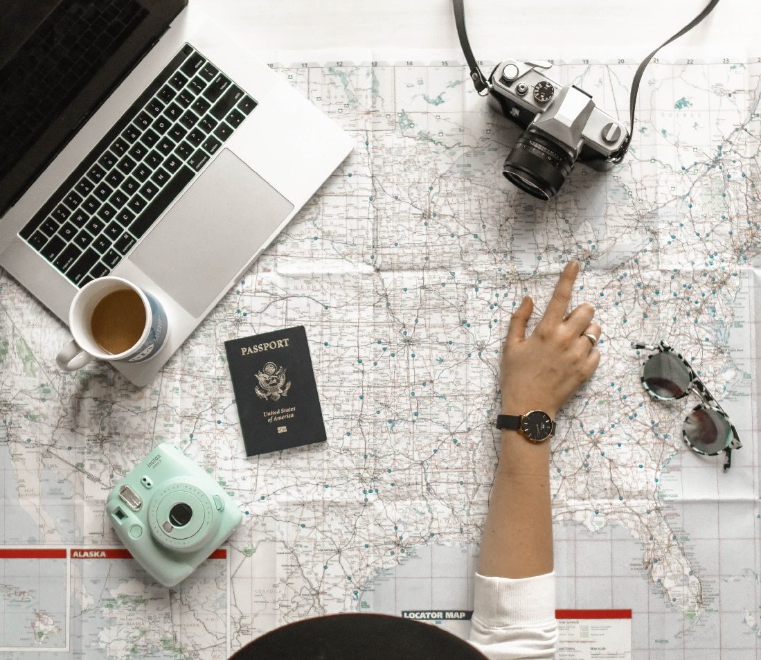 cameras, laptop, passport strewn across world map, with hand locating a destination